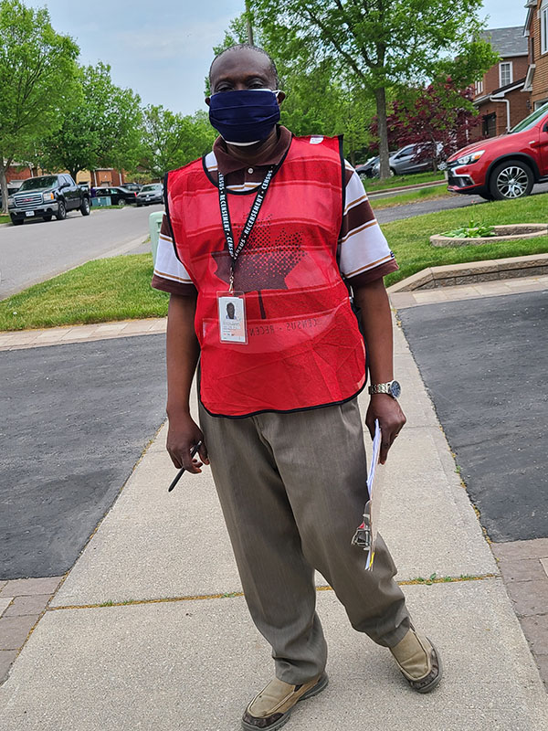 A Statistics Canada enumerator stands on the sidewalk holding a clipboard and pen, and wearing an a PPE mask as well as a Census 2021 red vest and an employee identification badge. 